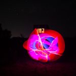 Glowing sphere of pink and blue light trails in front of a silhouetted WWII radar bunker at night