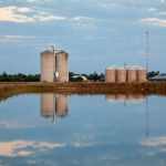 Grain silos reflecting on a still rural waterway under a bright sky